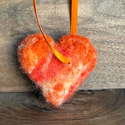 A felted heart in shades of orange with an orange ribbon attached for hanging by Lisa J. Byers